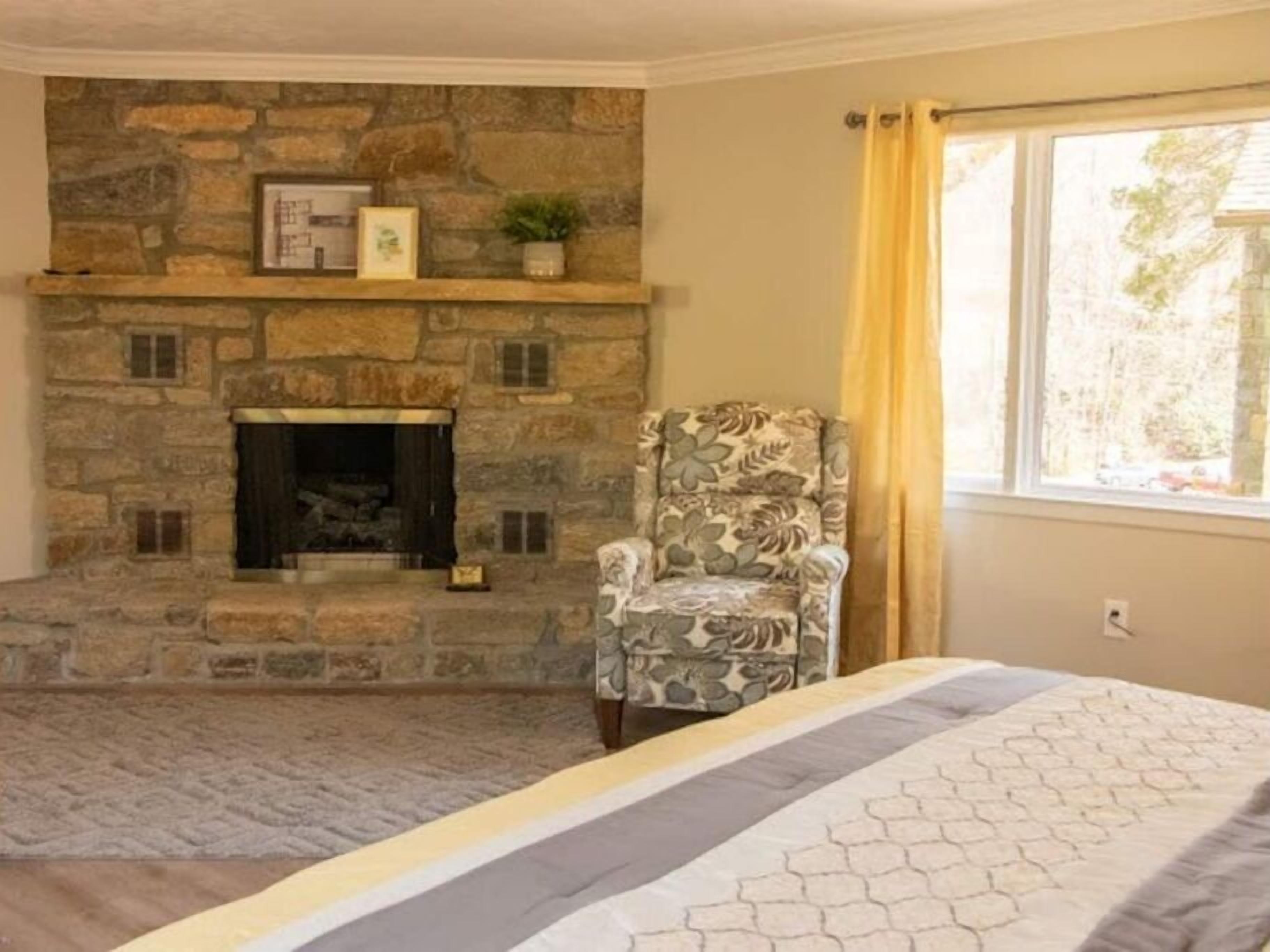 A room with a stone fireplace, a patterned armchair, and a bed with a geometric-patterned bedspread.