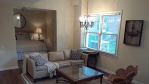 Um espaço compacto com sofá, mesa pequena, cadeira, cama, abajur, lustre e janela com persianas.