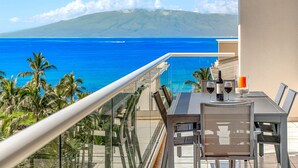 A balcony with a table, chairs, and a bottle of wine, overlooking a blue ocean and a mountain. - Maui Resort Rentals: Honua Kai Konea 649, Gorgeous 6th Floor 3BR w/ Stunning Ocean and Black Rock Views! (Lahaina)