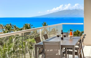 Un balcon avec une table dressée pour deux personnes, offrant une vue sur une mer bleue et limpide et sur une montagne.