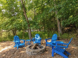 A fire pit with four blue Adirondack chairs arranged around it, set on a gravel area with trees in the background.
