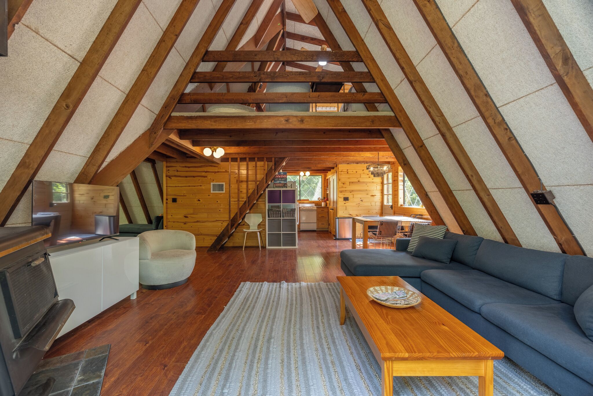 A cozy attic room with a wooden ceiling, a sofa, a coffee table, and a kitchen area with a stove and sink.