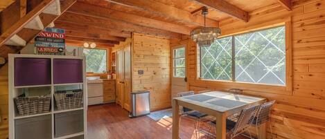 A wooden cabin interior with a dining table, chairs, a kitchen area, and a large window.