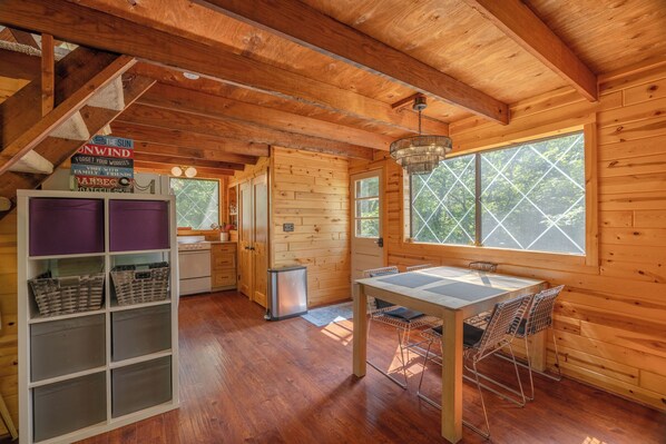 A wooden cabin interior with a dining table, chairs, a kitchen area, and a large window.