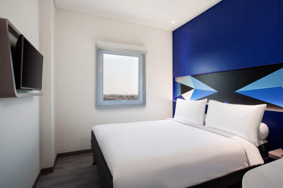 Une chambre d’hôtel moderne dotée d’un grand lit, d’un téléviseur à écran plat fixé au mur et d’une fenêtre offrant une vue sur le paysage urbain.
