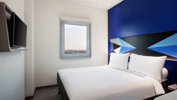 A modern hotel room with a large bed, a flat-screen TV mounted on the wall, and a window providing a view of the cityscape.