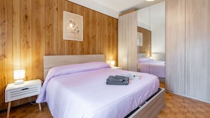 A bedroom with a wooden wall panel, a bed with a white headboard, a bedside table with a lamp, and a large mirror. - Maison Pro de Solari by Interhome (Fenis)