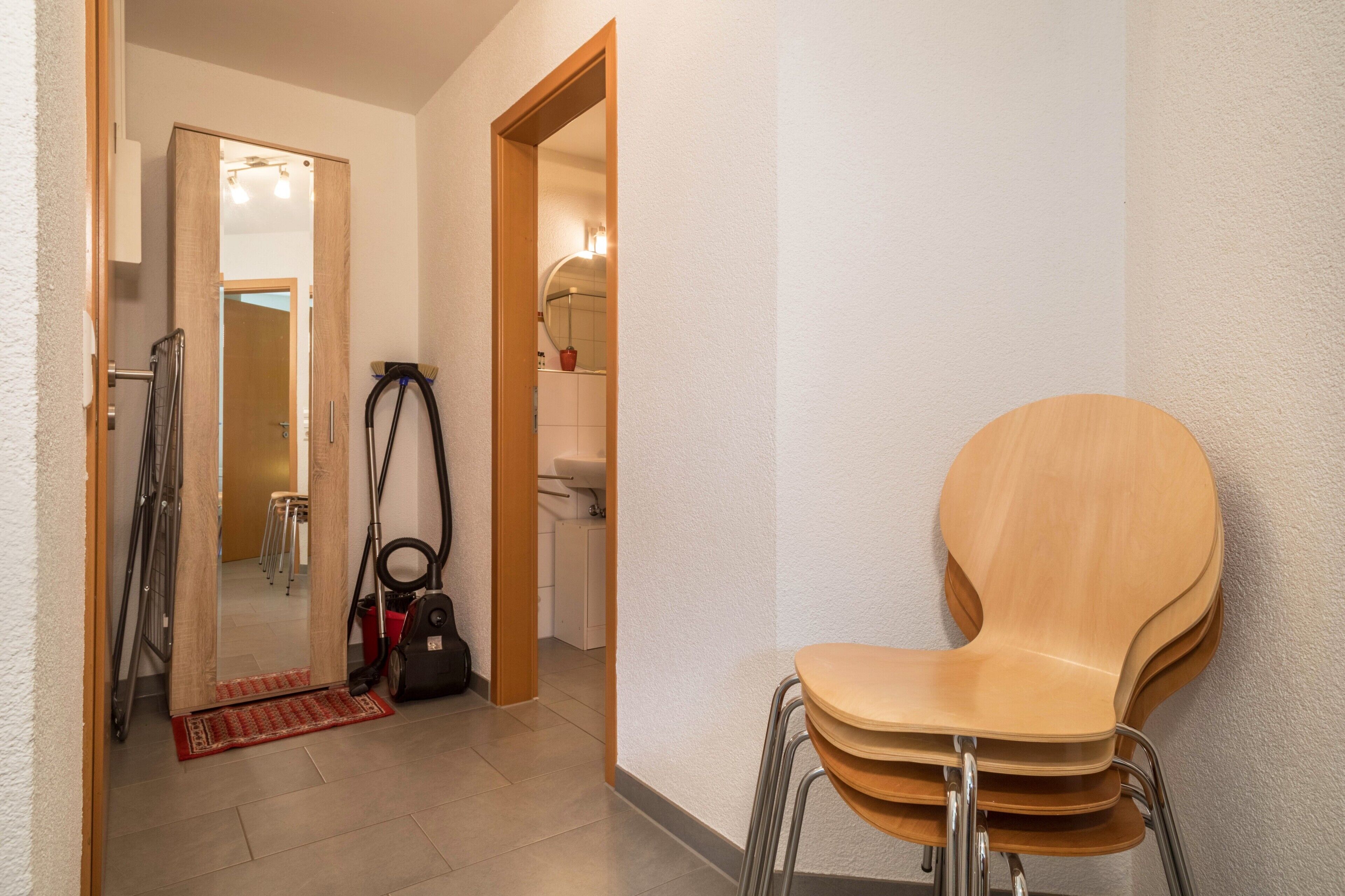 A hallway with a wooden chair, a vacuum cleaner, and a mirror reflecting a bathroom.