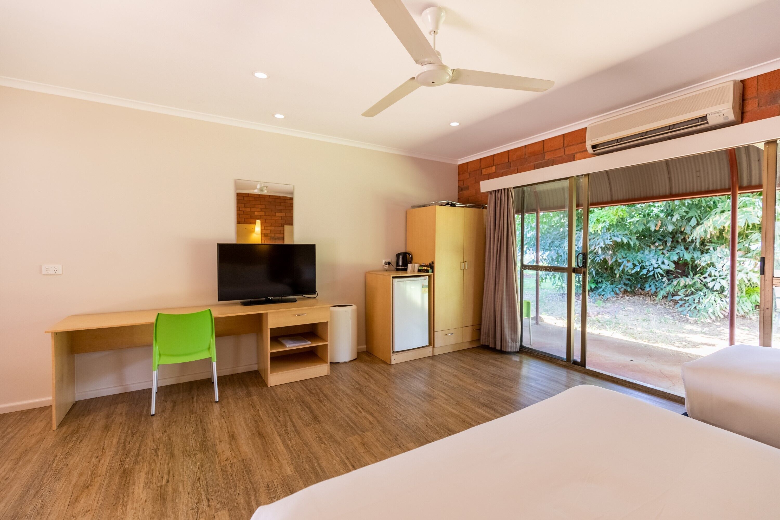 Habitación de hotel con escritorio de madera, una silla verde, cama, televisión, mini refrigerador y ventana grande con vistas a zonas verdes.