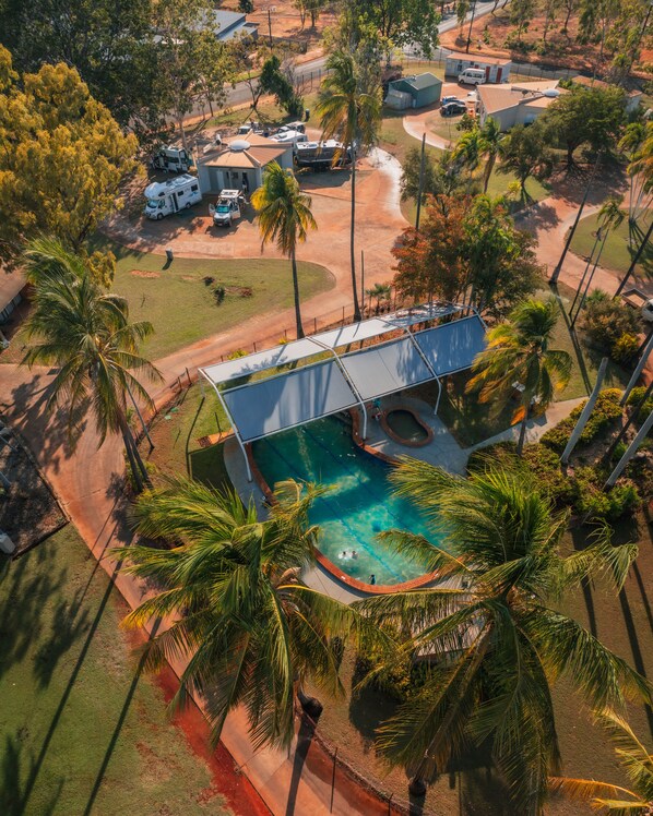 An aerial view of a residential area with a swimming pool surrounded by palm trees, a covered outdoor area, and RVs parked nearby. - Contour Hotel Katherine (Katherine East)