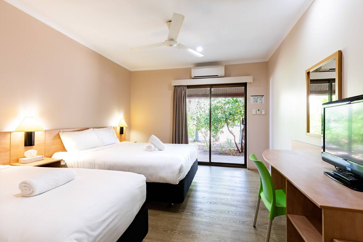 A hotel room with two beds, a desk with a computer, a green chair, and a ceiling fan.