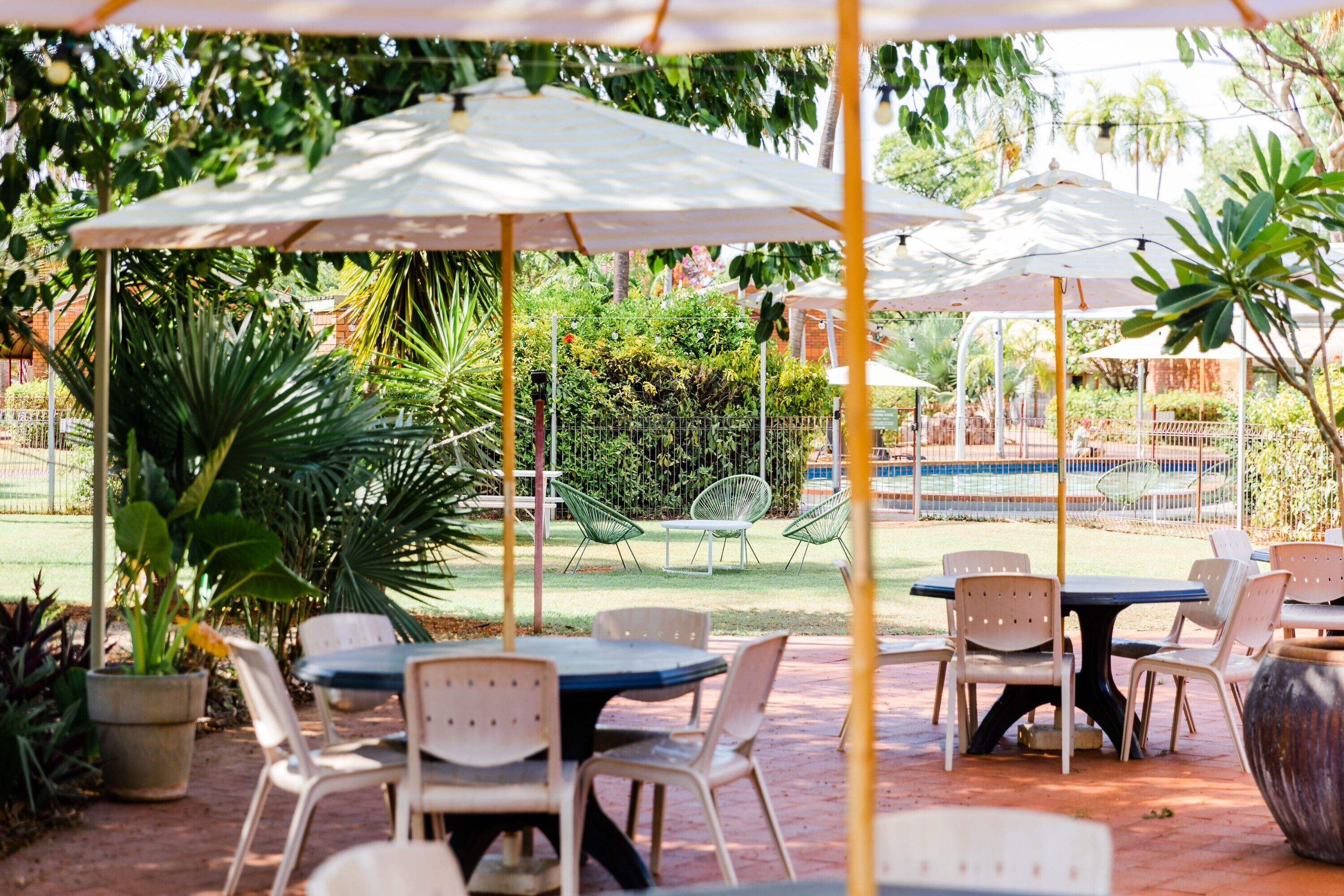 Un patio al aire libre con mesas y sillas bajo sombrillas, rodeado de follaje y una piscina al fondo.