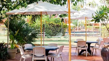 Un patio al aire libre con mesas y sillas bajo sombrillas, rodeado de follaje y una piscina al fondo.