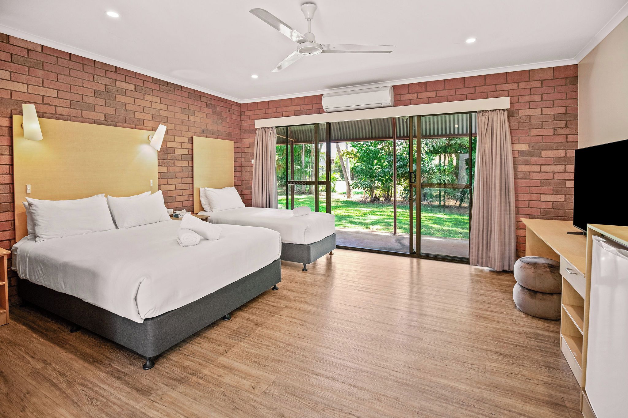 A hotel room with two beds, a ceiling fan, an air conditioning unit, a television, and a view of a garden through a glass door.