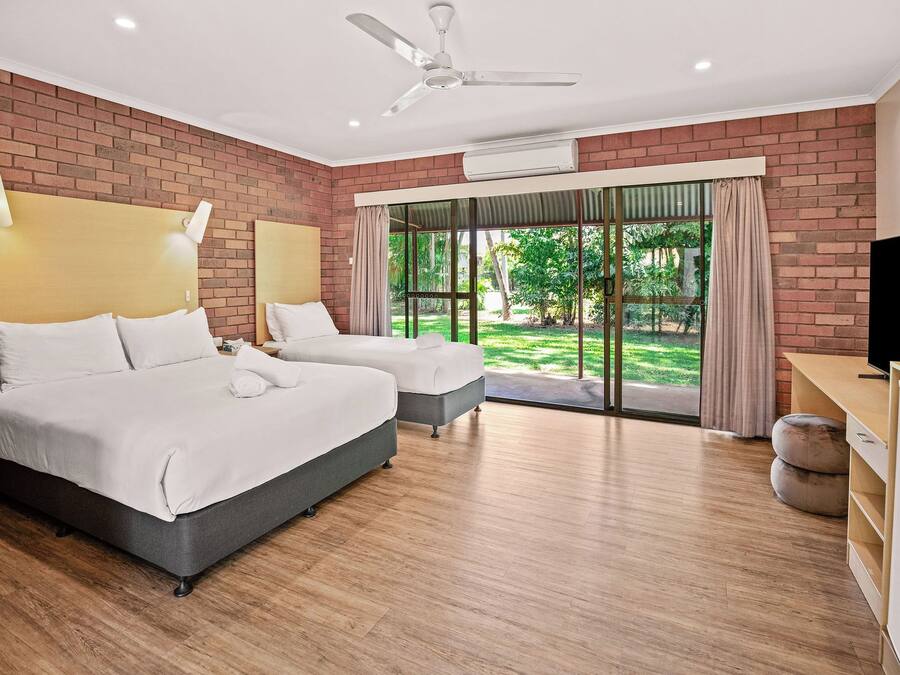 A hotel room with two beds, a ceiling fan, an air conditioning unit, a television, and a view of a garden through a glass door.