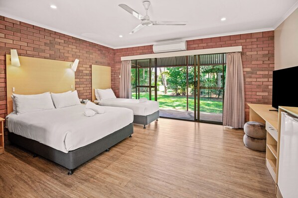 A hotel room with two beds, a ceiling fan, an air conditioning unit, a television, and a view of a garden through a glass door. - Contour Hotel Katherine (Katherine East)
