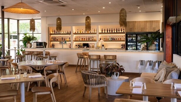 A restaurant interior with wooden tables and chairs, a bar area with shelves of bottles, and a large window providing natural light. - Contour Hotel Katherine (Katherine East)
