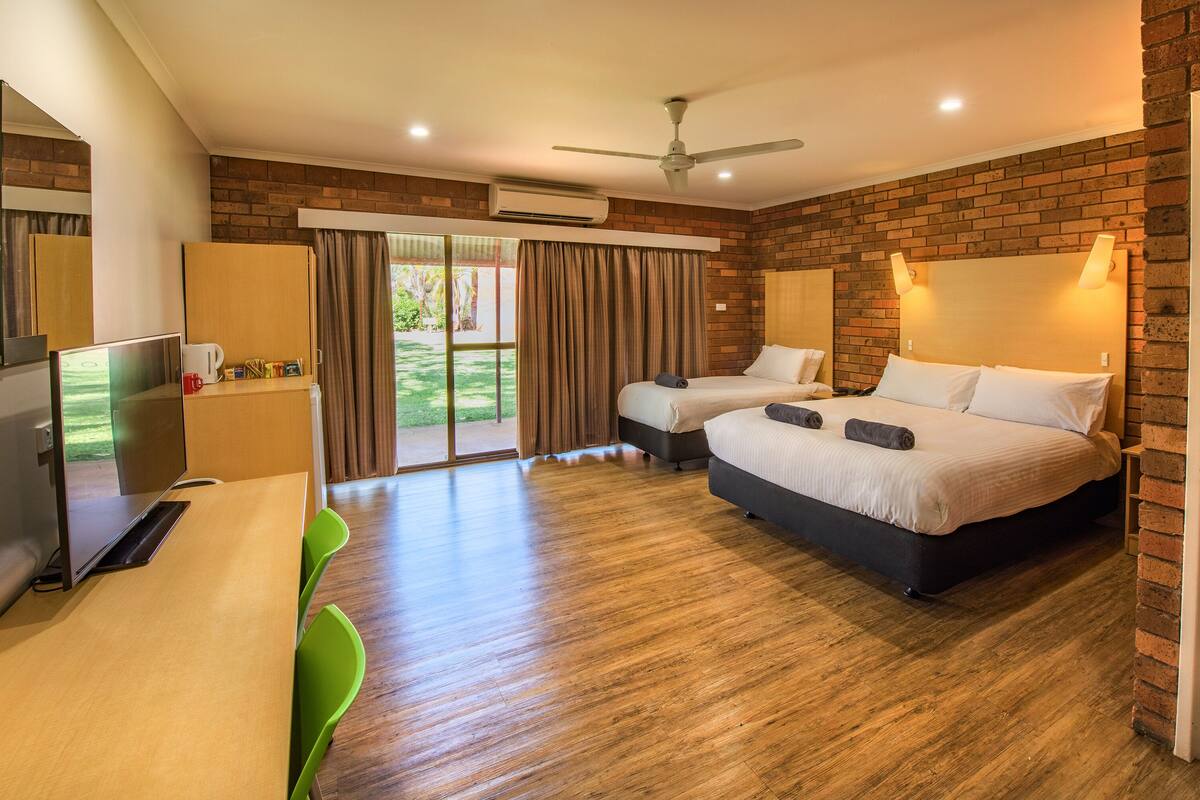 A modern hotel room with a brick wall, two beds, a desk, a TV, and a ceiling fan.