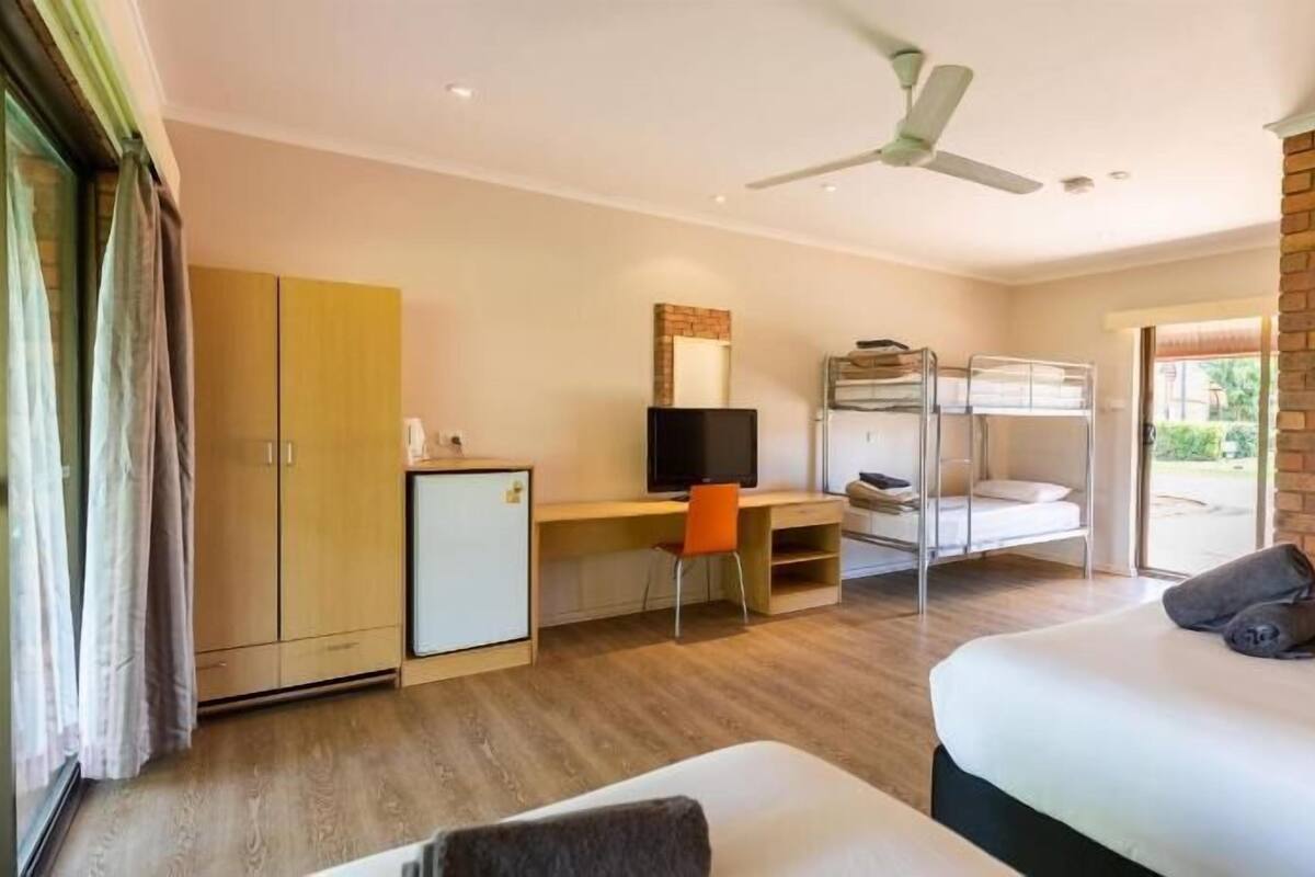 A room with a bunk bed, a desk with a computer, a small refrigerator, and a ceiling fan.