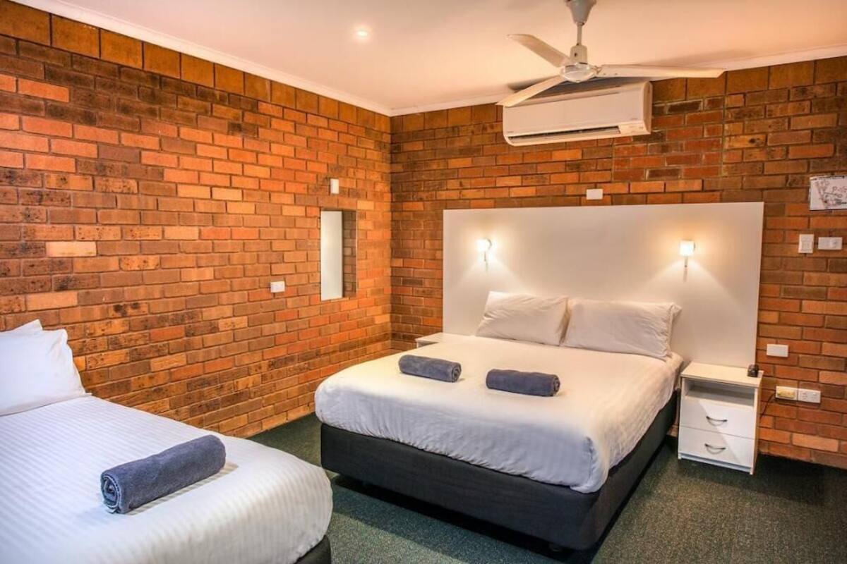 A hotel room with a brick wall, a large bed with white bedding, a nightstand, and a ceiling fan.
