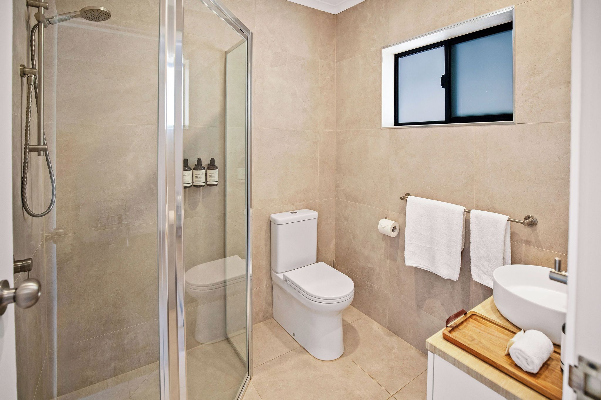 A modern bathroom with a glass-enclosed shower, a toilet, a sink, and a window above the mirror.