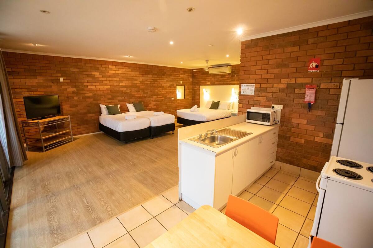 A compact hotel room with two beds, a kitchenette, a TV, and a brick wall.