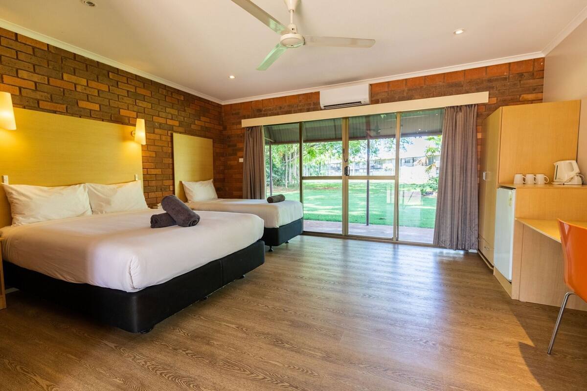 A hotel room with two beds, a desk, a chair, a wardrobe, and a sliding glass door leading to a garden.