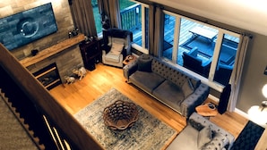 A living room with a fireplace, a sofa, a coffee table, and a large window offering a view of the deck. - Top Rated Waterfront Cottage + Sauna | Beach | Private Dock (Georgian Bluffs)