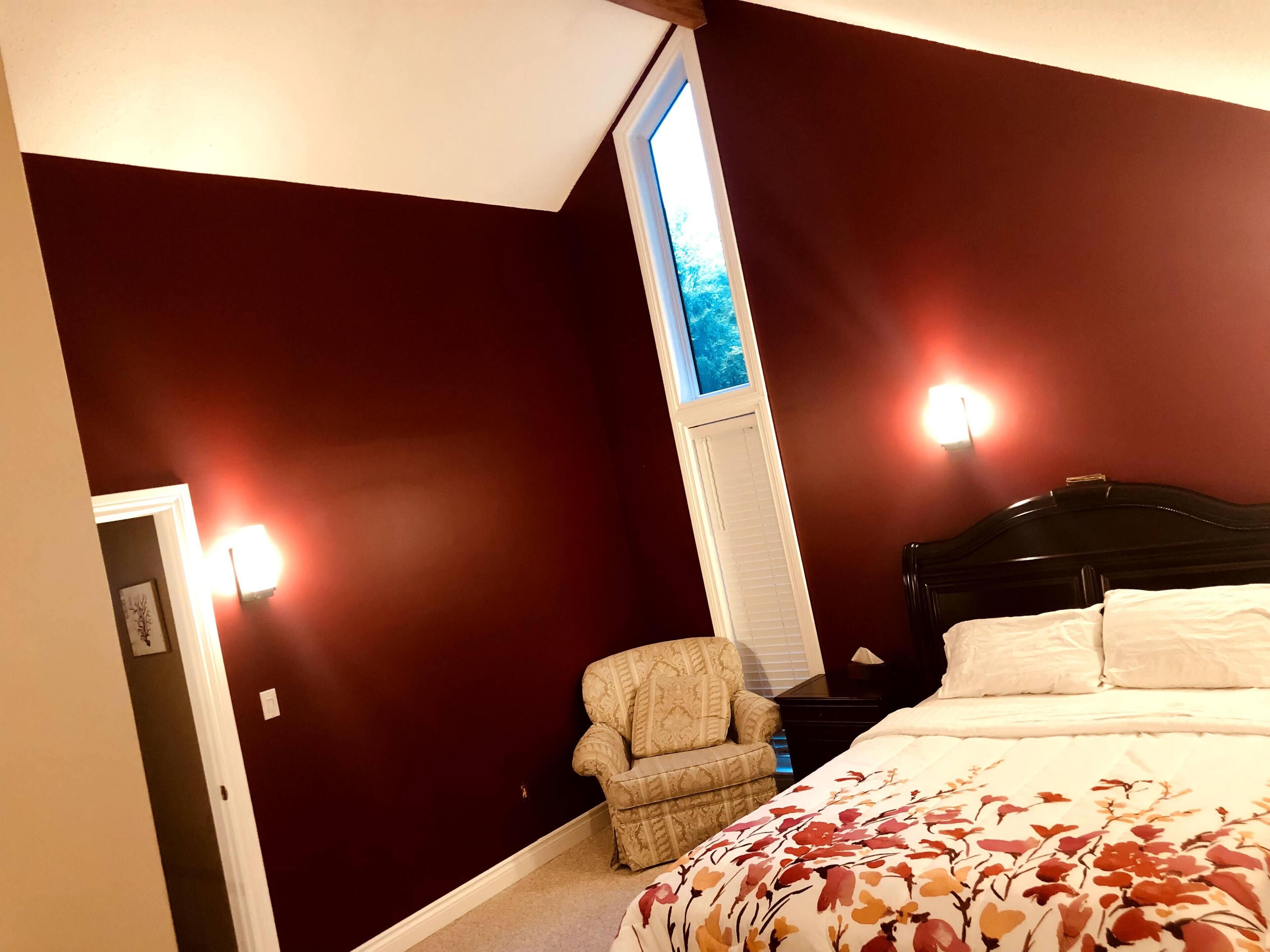 A bedroom with a dark red wall, a window with blinds, a bed with white bedding, a chair, and two wall-mounted lights.