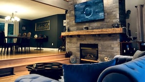 A modern living room with a fireplace, a dining area, and a large TV mounted on the wall. - Top Rated Waterfront Cottage + Sauna | Beach | Private Dock (Georgian Bluffs)