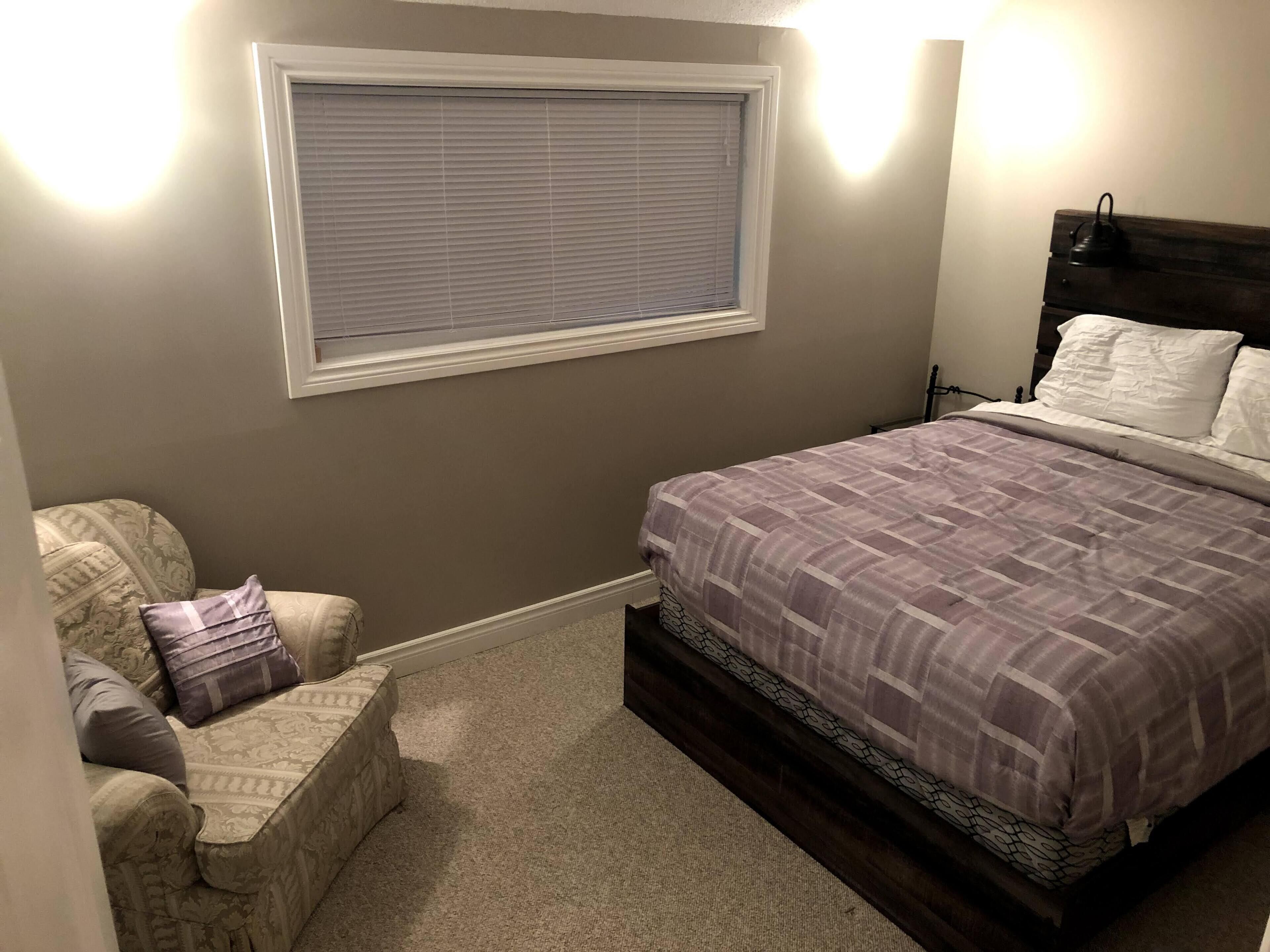 Een slaapkamer met een bed, een stoel, een raam met jaloezieën en een nachtkastje.