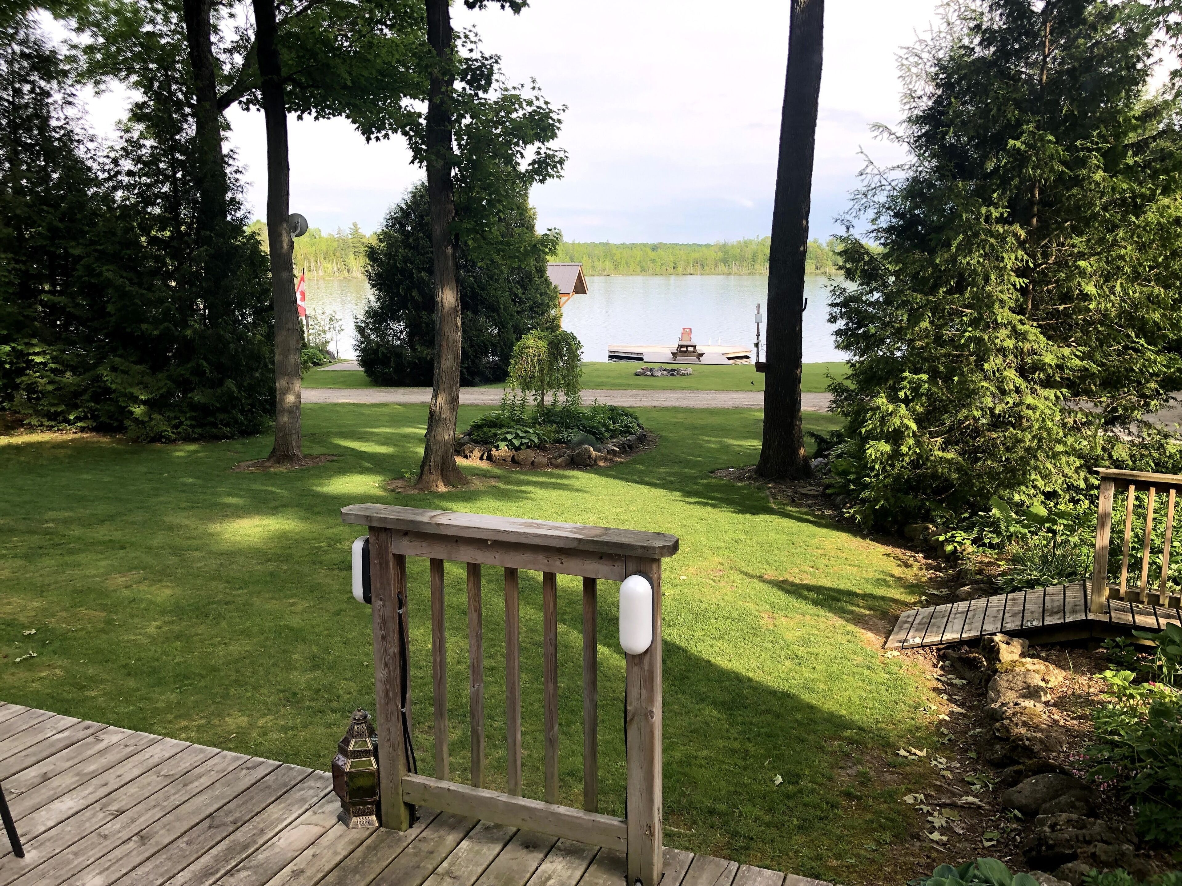 A wooden deck with a railing, a small lantern, and a view of a lake with a dock and a boat in the background.
