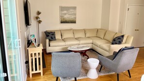 Una sala de estar moderna con un sofá seccional color beige, dos sillones grises, una mesita de noche de madera, un tablero de centro rojo y una lámpara de pie blanca.