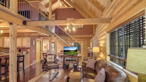A spacious living area with a high ceiling and wooden beams, a comfortable leather sofa, a television, and a large window with blinds.