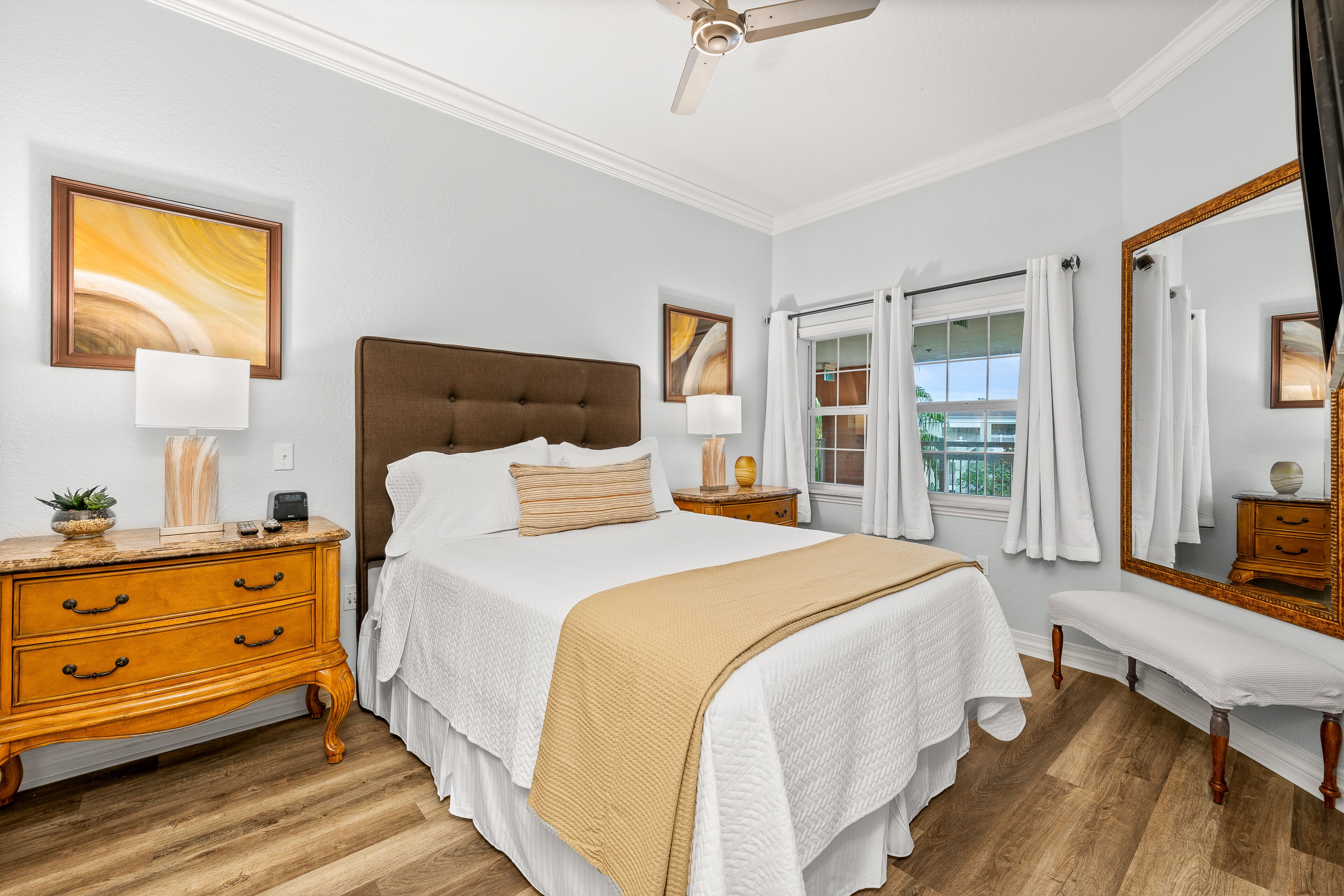 A bedroom with a large bed, wooden nightstands, a bench, a ceiling fan, and a window with curtains.
