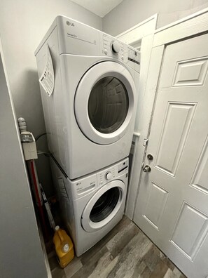 A laundry room with stacked washer and dryer units, a door, and a yellow detergent bottle on the floor. - Cedar Brook Manor - Rutgers | NYC | Jersey Shore - Union County - EXTENDED STAY (Plainfield)