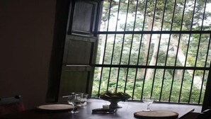 Un comedor con una mesa preparada para dos personas, un florero con flores y vistas a zonas verdes a través de ventanas con barrotes.