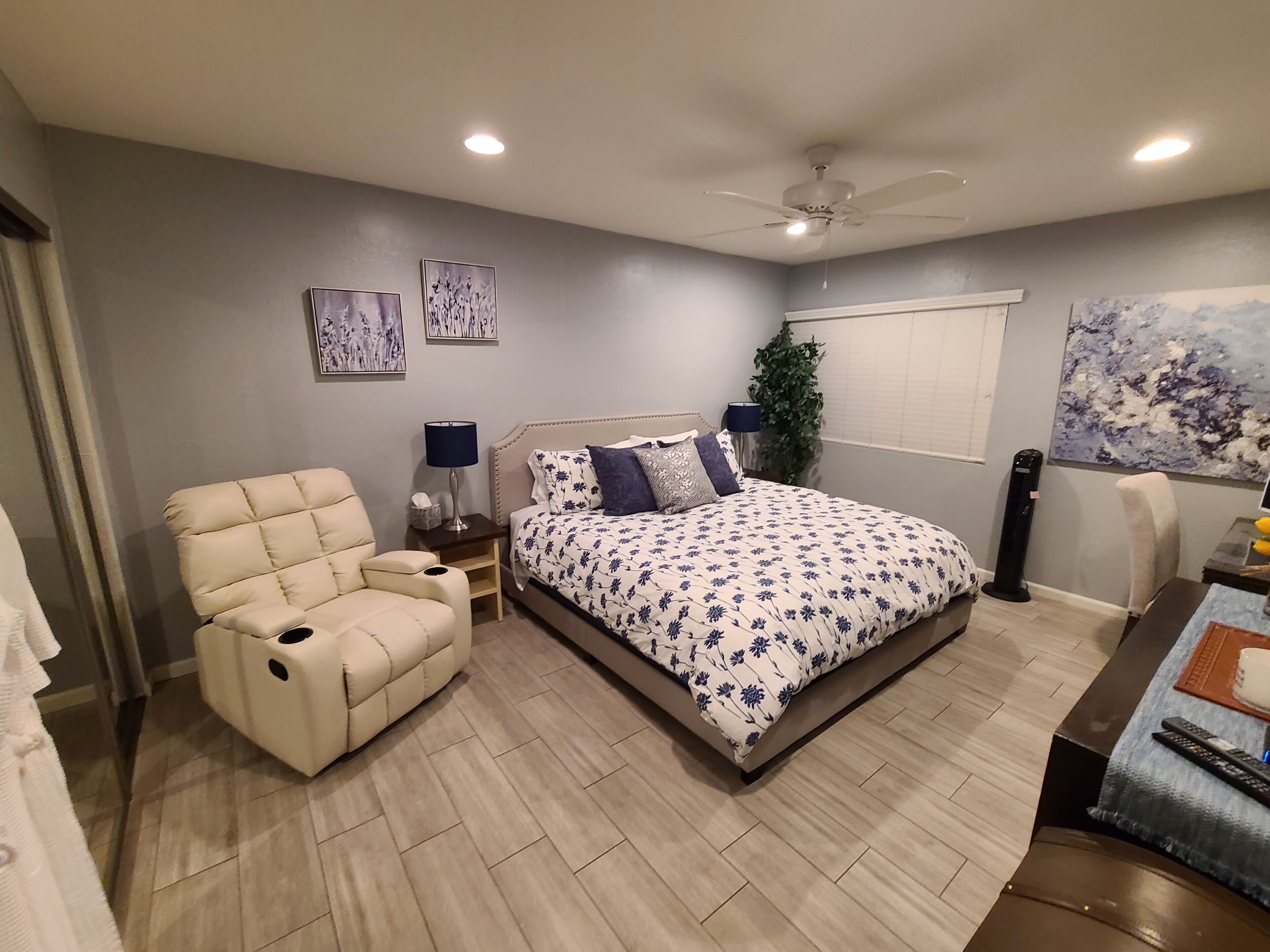 A bedroom with a bed, a chair, a nightstand, a lamp, a plant, and artwork on the walls.