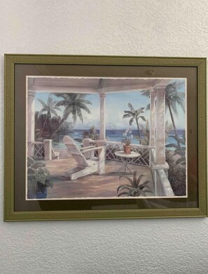 A framed painting of a tropical outdoor seating area with palm trees, a pool, and a view of the ocean.