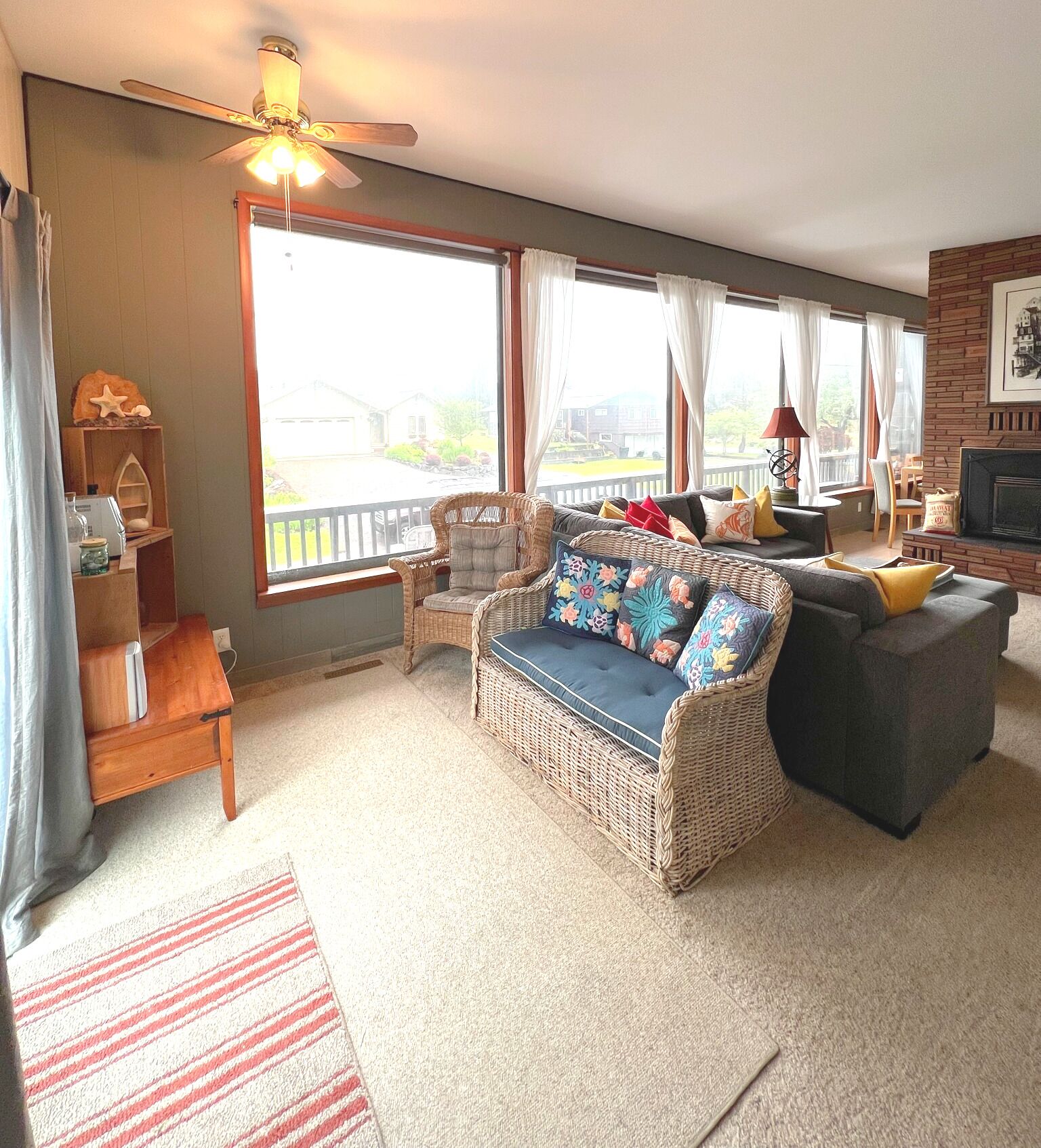 A cozy living room with a wicker sofa, a dark armchair, and a fireplace. There is a large window with curtains, a ceiling fan, and a rug on the floor.
