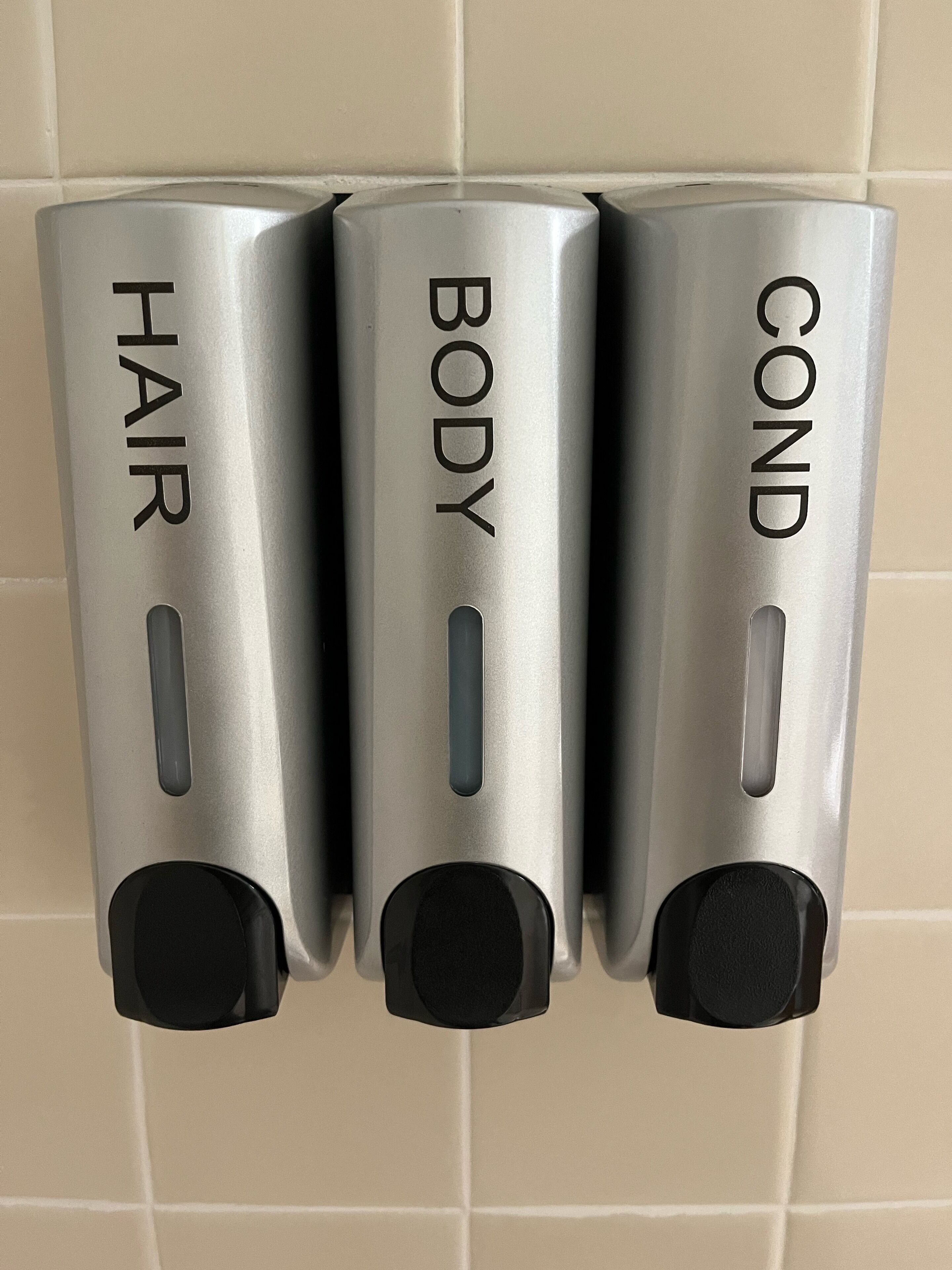 Three metallic dispensers labeled "HAIR," "BODY," and "COND" mounted on a tiled wall.