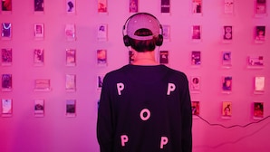 Une personne portant un pull foncé avec le mot « POP » écrit dans le dos, se tenant devant un mur orné de plusieurs tableaux encadrés.
