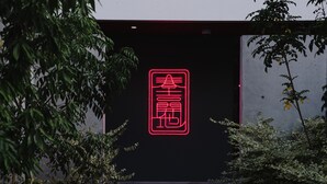 Une enseigne au néon avec des caractères chinois, fixée sur un mur sombre et entourée de verdure.