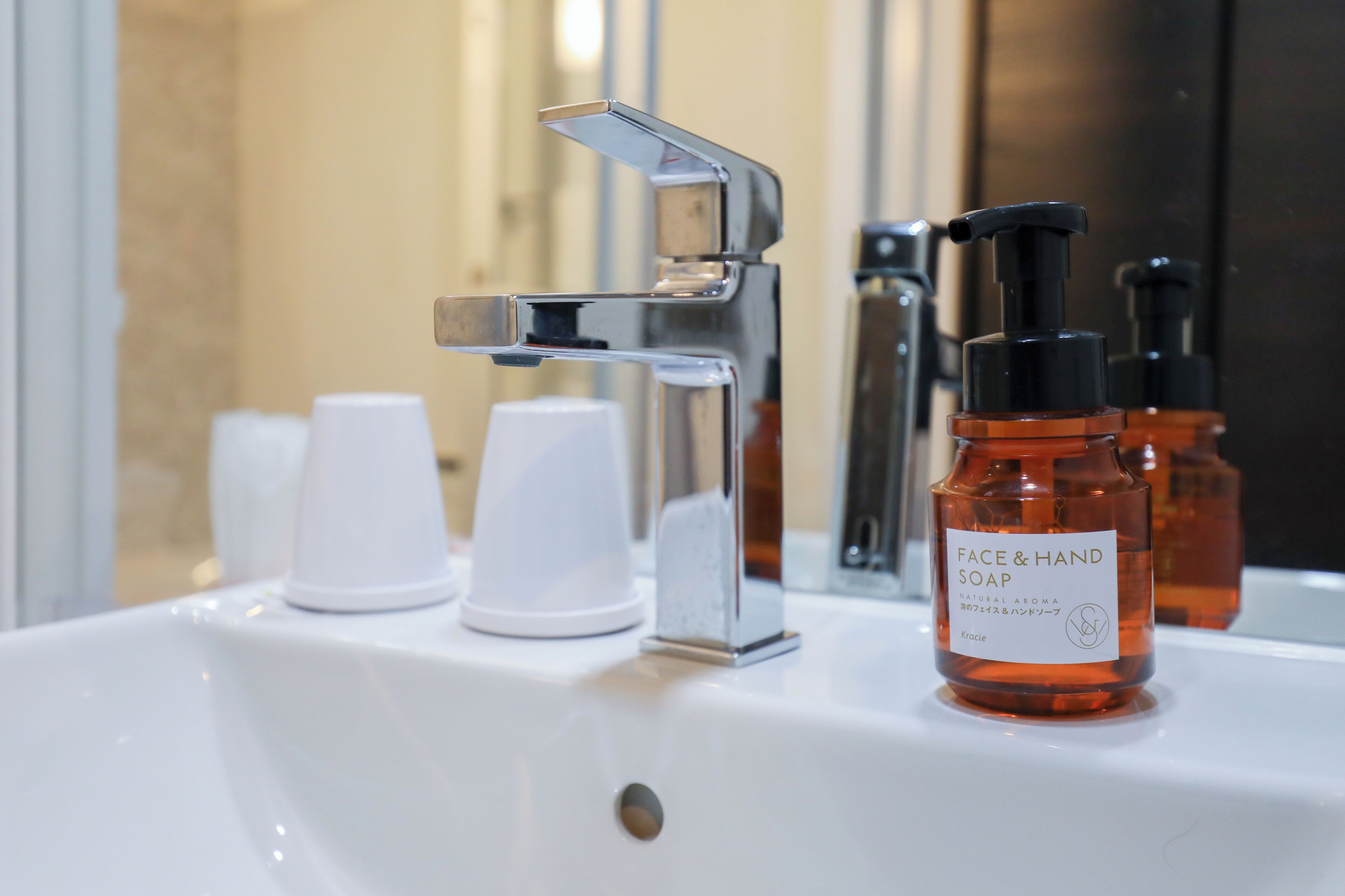 A bathroom sink with a chrome faucet, a bottle of face and hand soap, and two white paper cups.