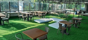 An outdoor seating area with tables and chairs, surrounded by large windows and greenery.