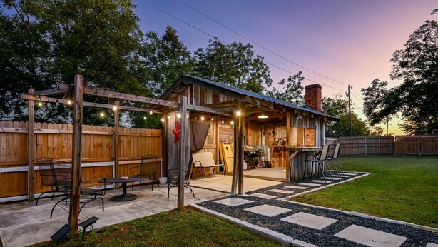 Un patio rústico al aire libre con una pérgola de madera, zona de estar y una pequeña construcción cubierta con guirnaldas de luces.