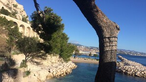A coastal landscape with a rocky shoreline, a tree, and a distant view of a town with buildings and a bridge. - Studio Avec Jardin (Marseille)