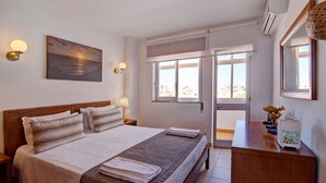 A hotel room with a bed, bedside lamps, a painting, a desk with a lamp, and a view through the window. - Apartment Vista Mar 4ºE on the Beach with Sea View, A/C & Balcony (Armação de Pêra)