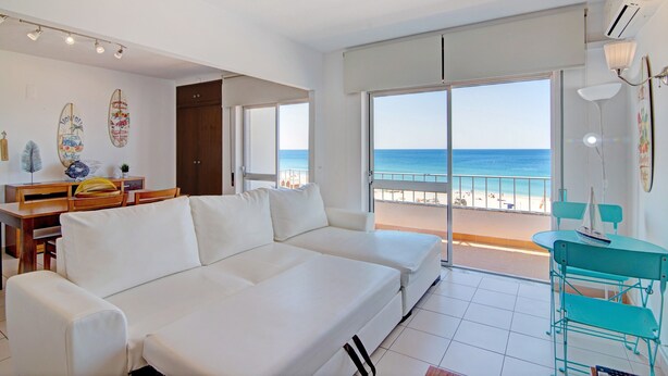 A modern living room with a white sofa, a dining table, and a balcony with a view of the beach. - Apartment Vista Mar 4ºE on the Beach with Sea View, A/C & Balcony (Armação de Pêra)