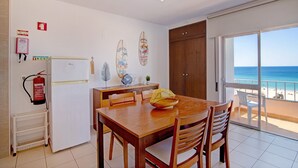 A dining area with a wooden table and chairs, a fire extinguisher, a refrigerator, and a view of the beach through a sliding glass door. - Apartment Vista Mar 4ºE on the Beach with Sea View, A/C & Balcony (Armação de Pêra)